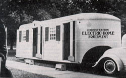 Photo of a 1937 trailer hitiched to the back of a car. The trailer is marked with the words Electrification Electric Home equipment.