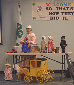 Young girl in pink dress with four dolls on a table and one doll in the floor with a horse and buggy.