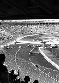 Black and white photo of cars racing with a large crowd.