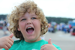 Young white boy with curly blond hair, smiling with mouth wide open and thumbs up.