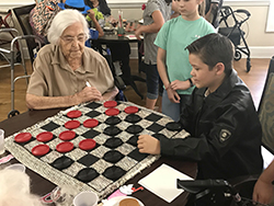 Two young boys, one in a black leather jacket, play checkers with older lady in retirement home.