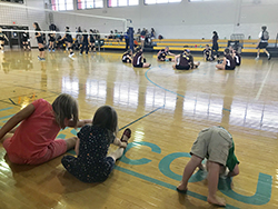Three kids stretchering in gym while vollyball players in background are also stretching.