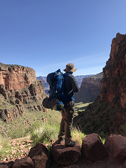 Man with hiking backpack and hat looks out on the landscape. Mountains in background.