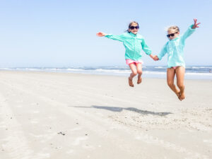 2 girls in sunglasses on beach holding hands and jumping in the air