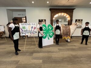 Nash County 4-H Carolina Choppers wear aprons and pose by challenge sign and 4-H clover logo